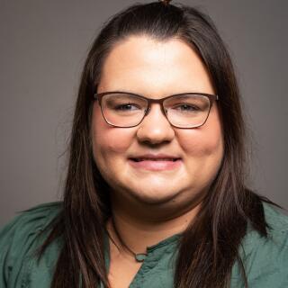 a headshot of a woman wearing a green top with long dark hair and glasses